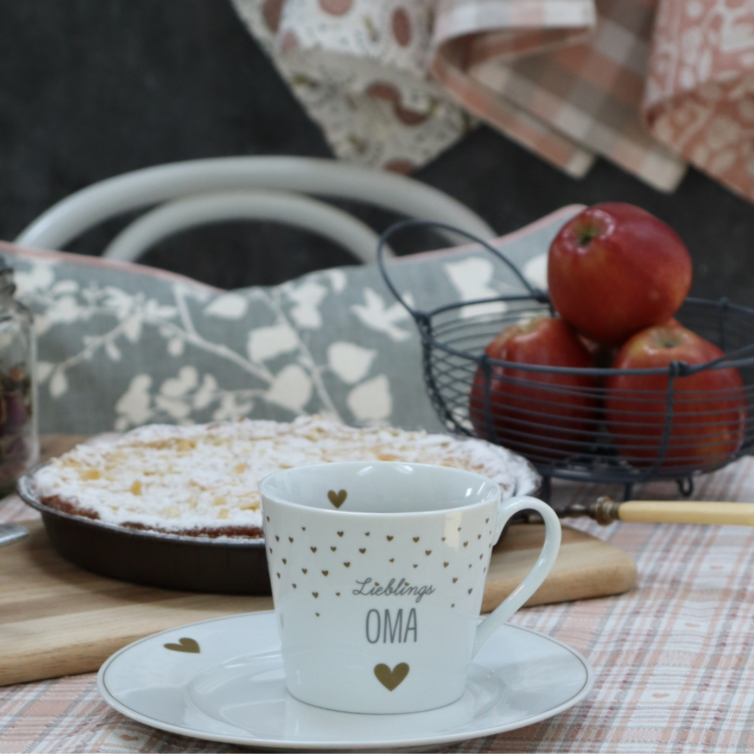 Frühstückstisch gedeckt mit Teller und der Tasse "Lieblingsoma" im Hintergrund Äpfel im Landhauskorb und frischer Kuchen.